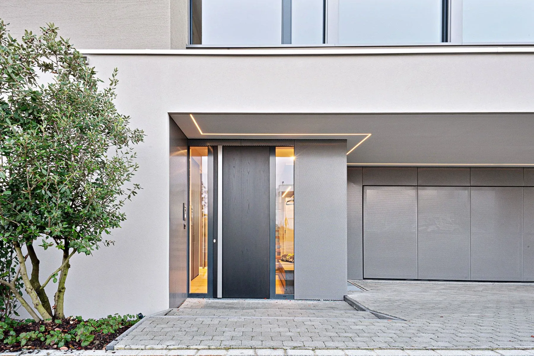 Aussenansicht auf die mit Holz beplankte Haustüre, den Eingangsbereich mit Lochblechverkleidung und integriertem Garagentor