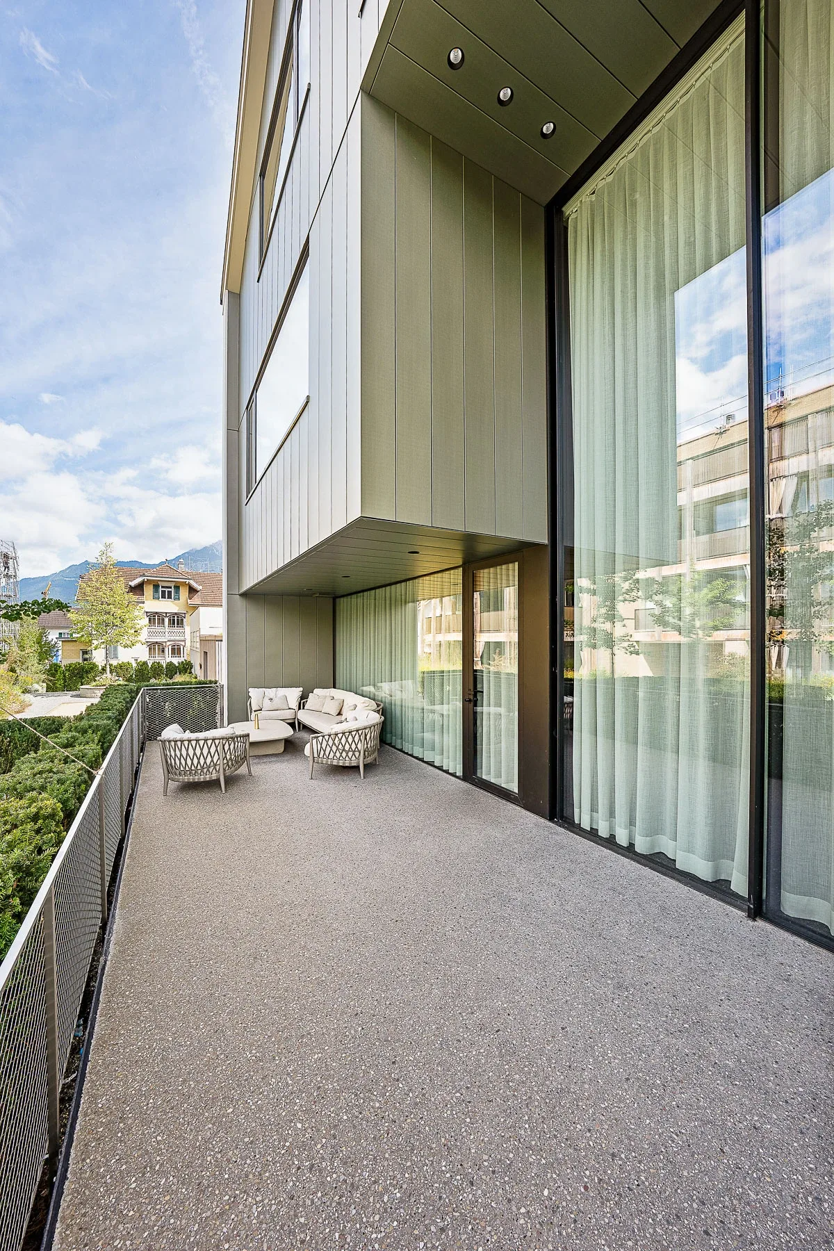 Balkon der Loftwohnung mit raumhoher Sky-Frame Verglasung, Vorhaengen und weissen Lounge-Sesseln