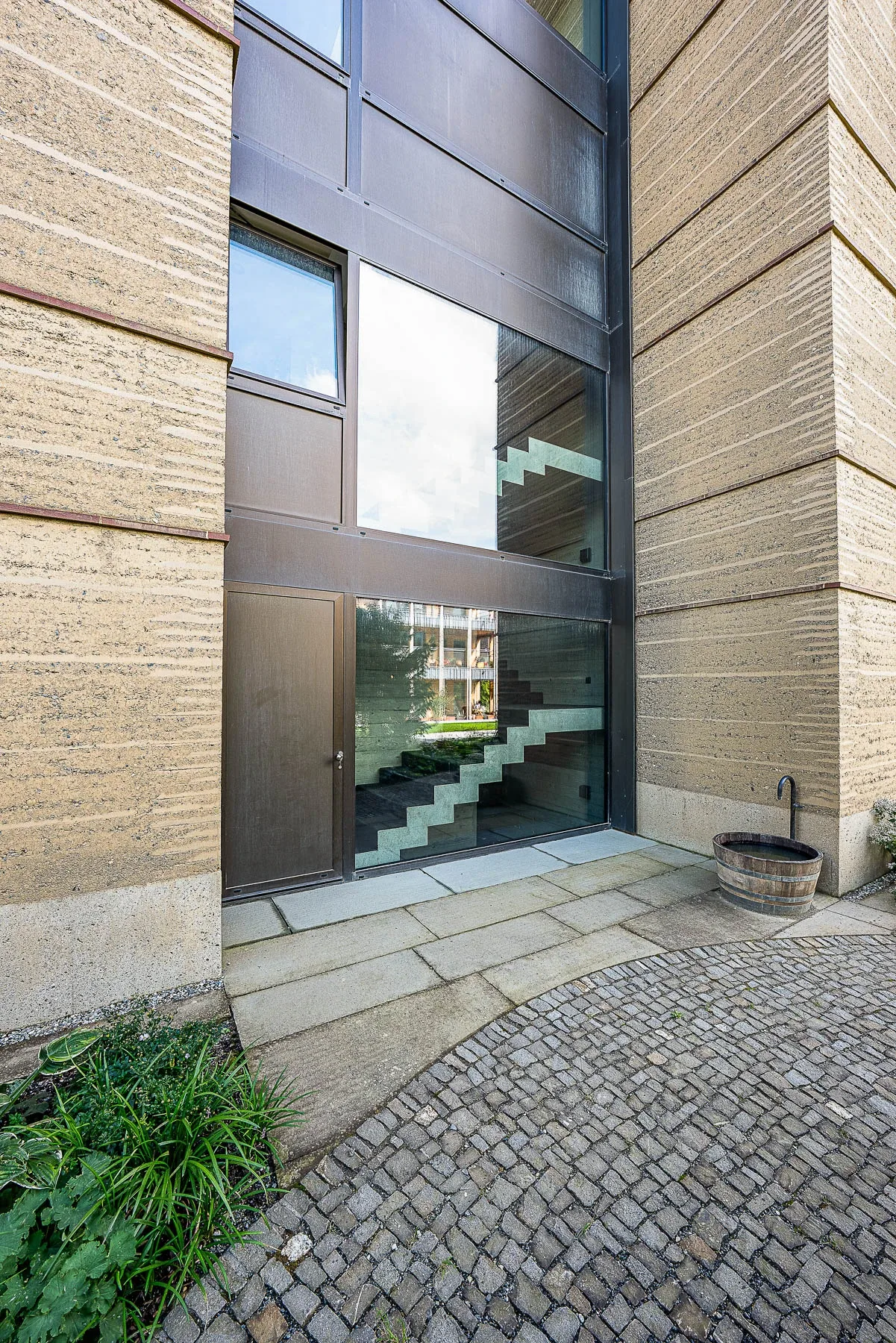 Nahaufnahme Eingang zwischen Stampflehm-Waenden mit verglastem Treppenhaus