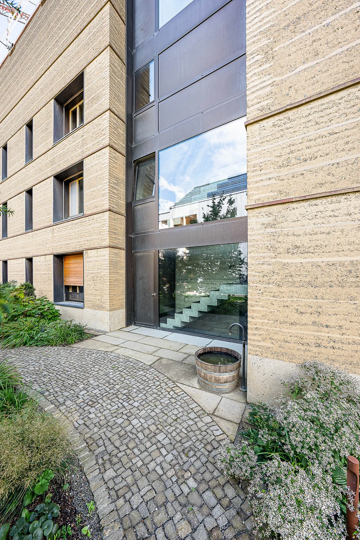 Eingangsbereich Haus der Freunde zwischen Stampflehm-Fassaden mit verglastem Treppenhaus und Kopfsteinpflaster