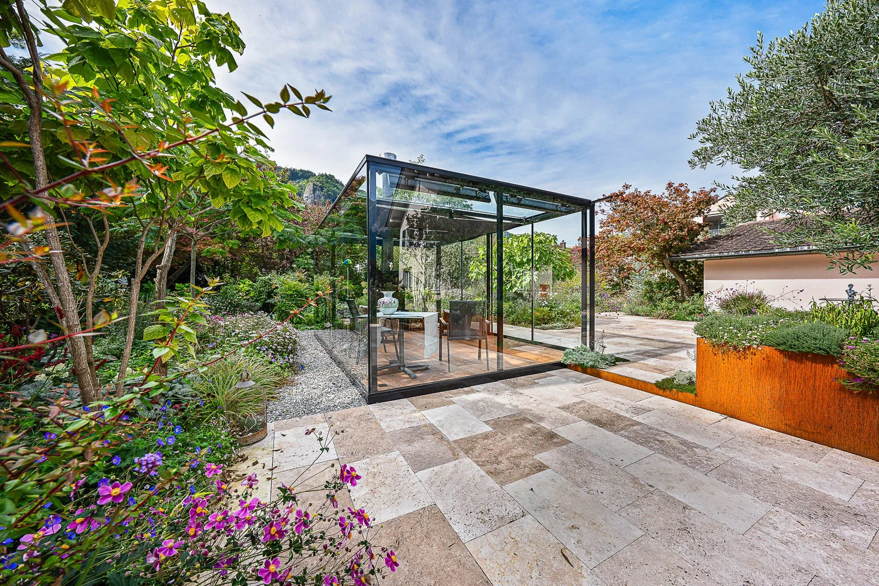 Schraege Gesamtansicht der Glaslounge mit Natursteinplatten und bluehendem Garten