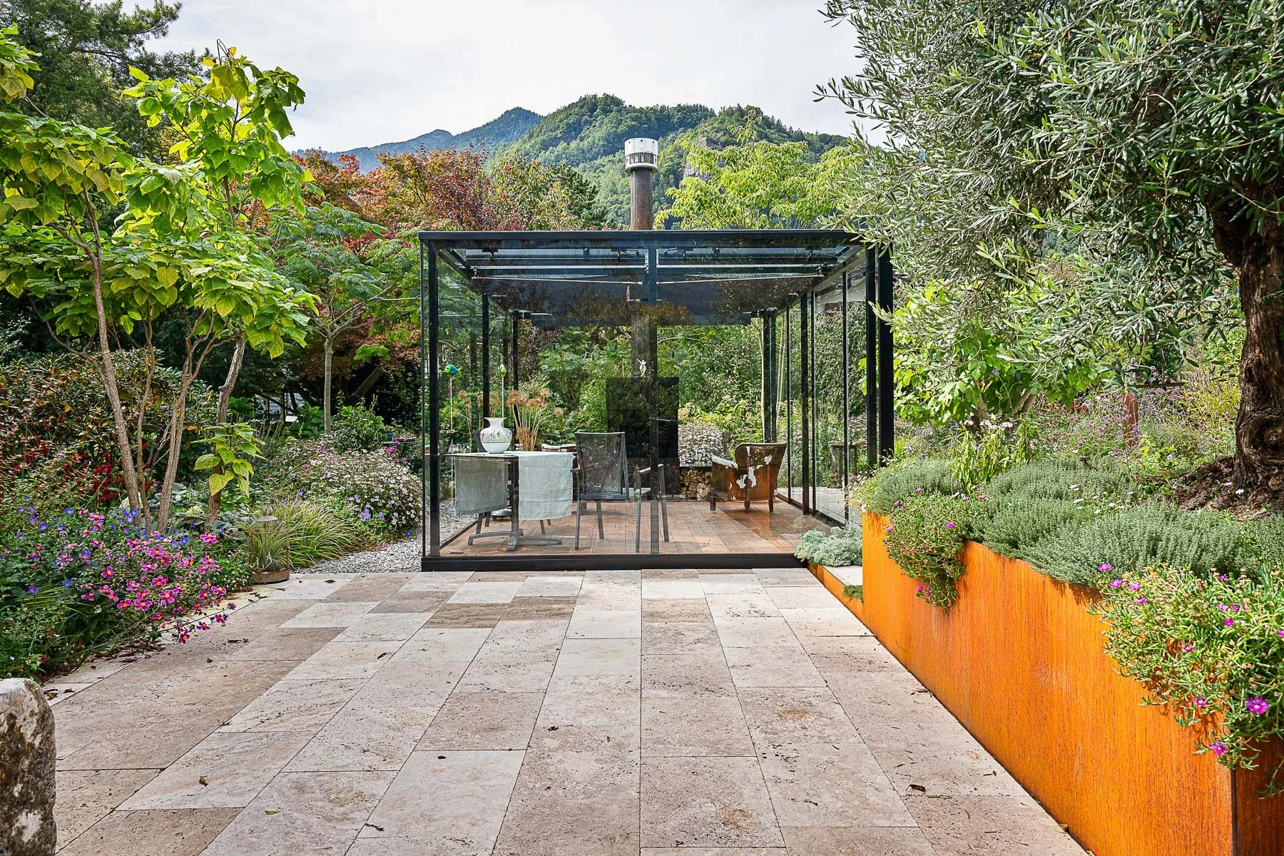 Outdoor Cigar Lounge frontal mit Natursteinplatten-Terrasse, Corten-Pflanztrog und Bergkulisse