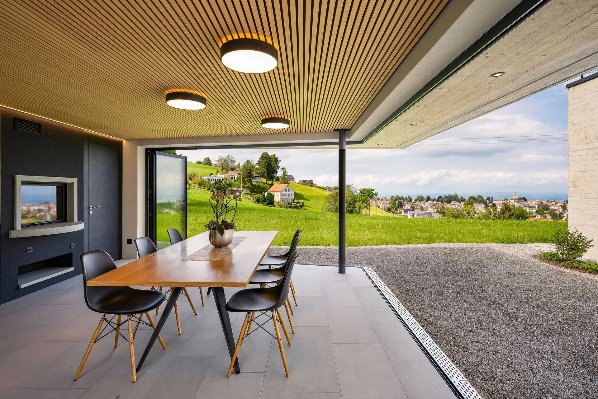 Innenansicht Pavillon mit Esstisch, offener Verglasung und Blick auf gruene Wiese und Dorf