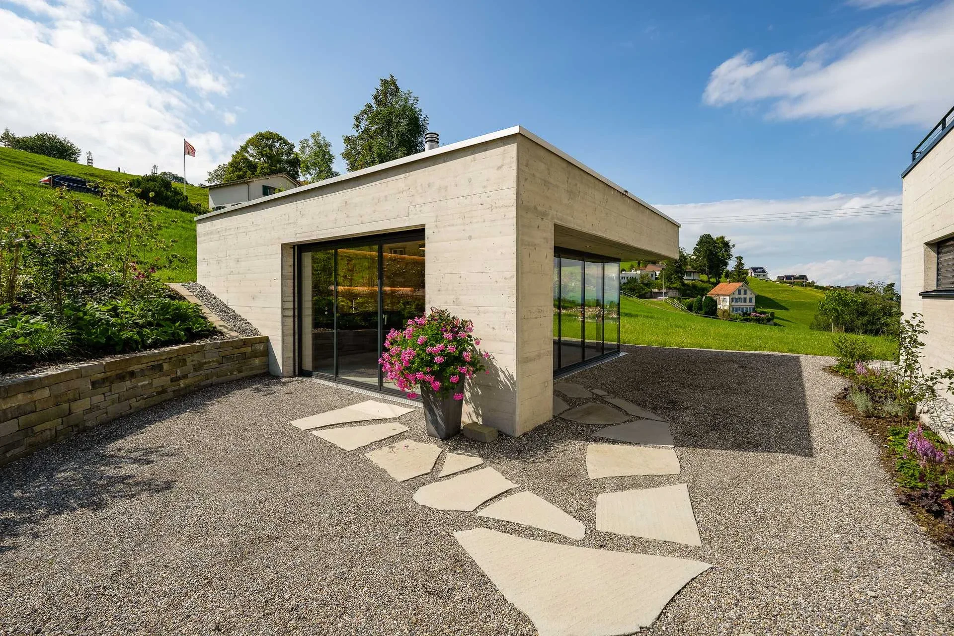 Pavillon-Eingang frontal mit Sichtbeton-Fassade und Blumentopf auf Naturstein-Trittplatten