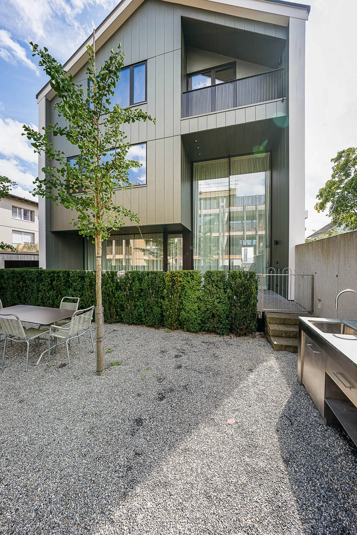 Erdgeschoss-Gartenbereich mit Metallverkleidung-Fassade, Kiesplatz, Edelstahl-Aussenkueche und jungem Baum